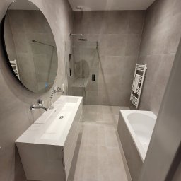 Firma Produkcyjno-Usługowa Przemysław Moskała - Nowoczesna łazienka w odcieniach szarości z owalnym lustrem, białą umywalką nablatową z szafką, kabiną prysznicową typu walk-in i grzejnikiem drabinkowym z ręcznikiem.