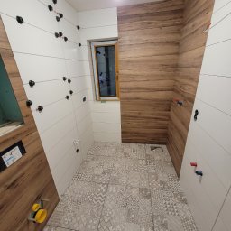 Firma Produkcyjno-Usługowa Przemysław Moskała - Łazienka w trakcie wykańczania: białe i drewnopodobne płytki ścienne ułożone naprzemiennie, wzorzysta szara podłoga, widoczne elementy instalacji wodnej i uchwyty do płytek na ścianach.