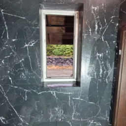 Łazienka po remoncie: czarne płytki imitujące marmur na ścianie z oknem, widok na zewnątrz. Białe fugi i biała rama okienna. W tle żarówka na kablu.