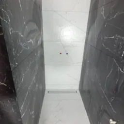 Wnętrze łazienki z czarno-białymi płytkami imitującymi marmur, ułożonymi wzdłuż ścian i na podłodze. Widoczny odpływ liniowy i wystające rurki.