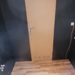 Jasne panele podłogowe z drewnianym wzorem, ściany pomalowane na ciemny kolor, widoczna drewniana futryna drzwiowa z metalową klamką i gniazdko elektryczne.