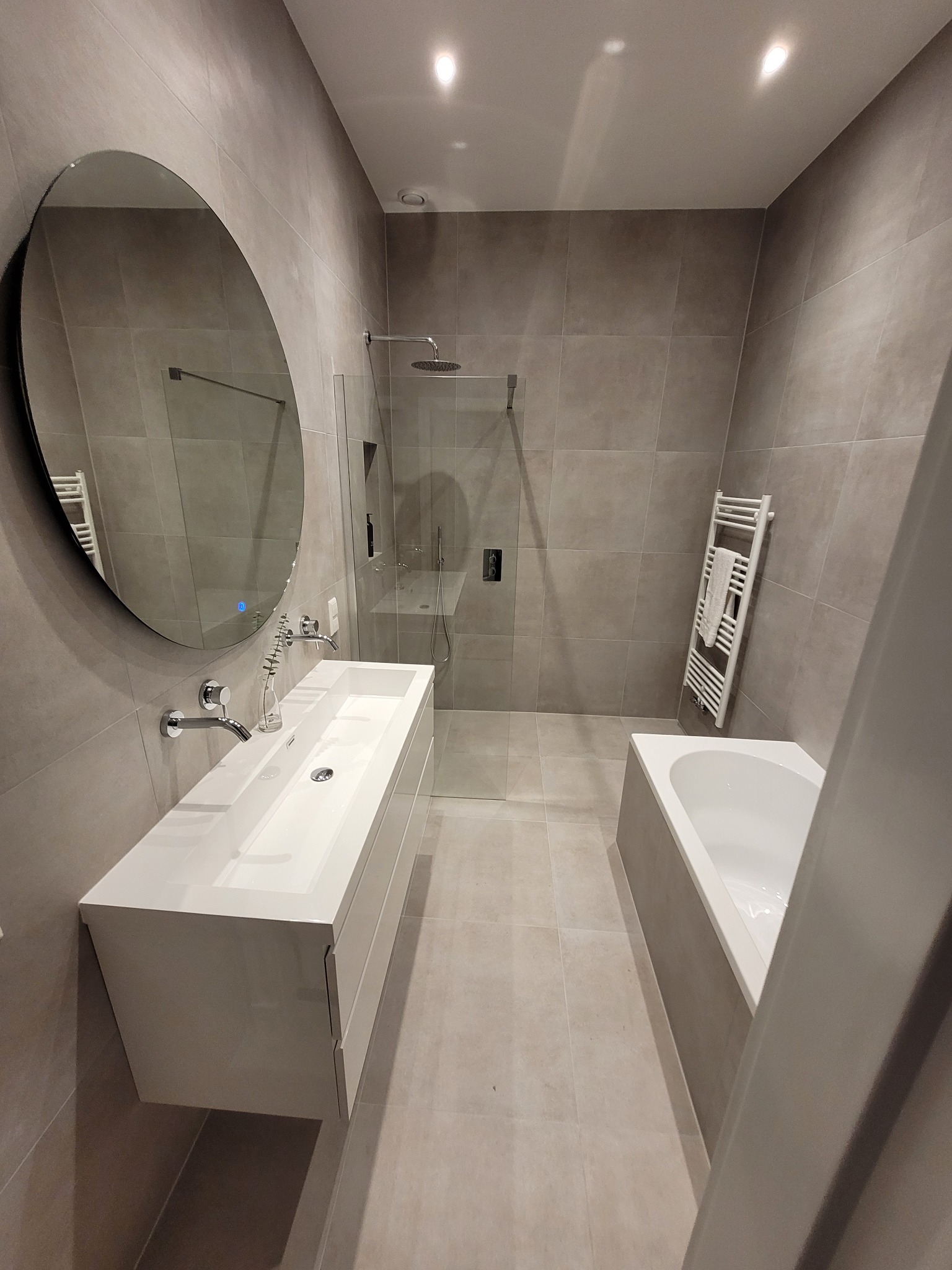 Nowoczesna łazienka w odcieniach szarości z owalnym lustrem, białą umywalką nablatową z szafką, kabiną prysznicową typu walk-in i grzejnikiem drabinkowym z ręcznikiem.