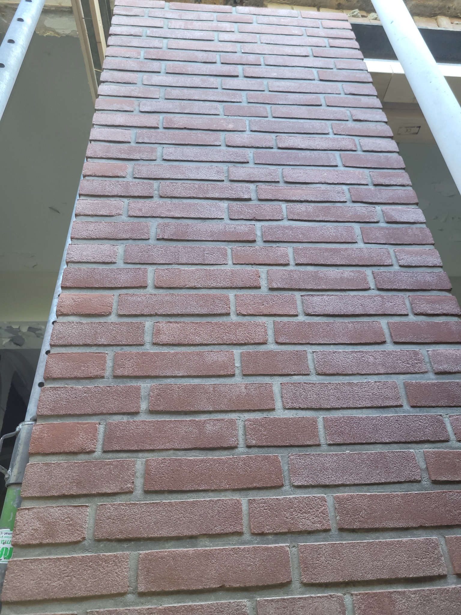 Wykończony komin obłożony cegłą klinkierową w odcieniach czerwieni i brązu, widoczny z dołu, z fragmentami rusztowania po bokach.