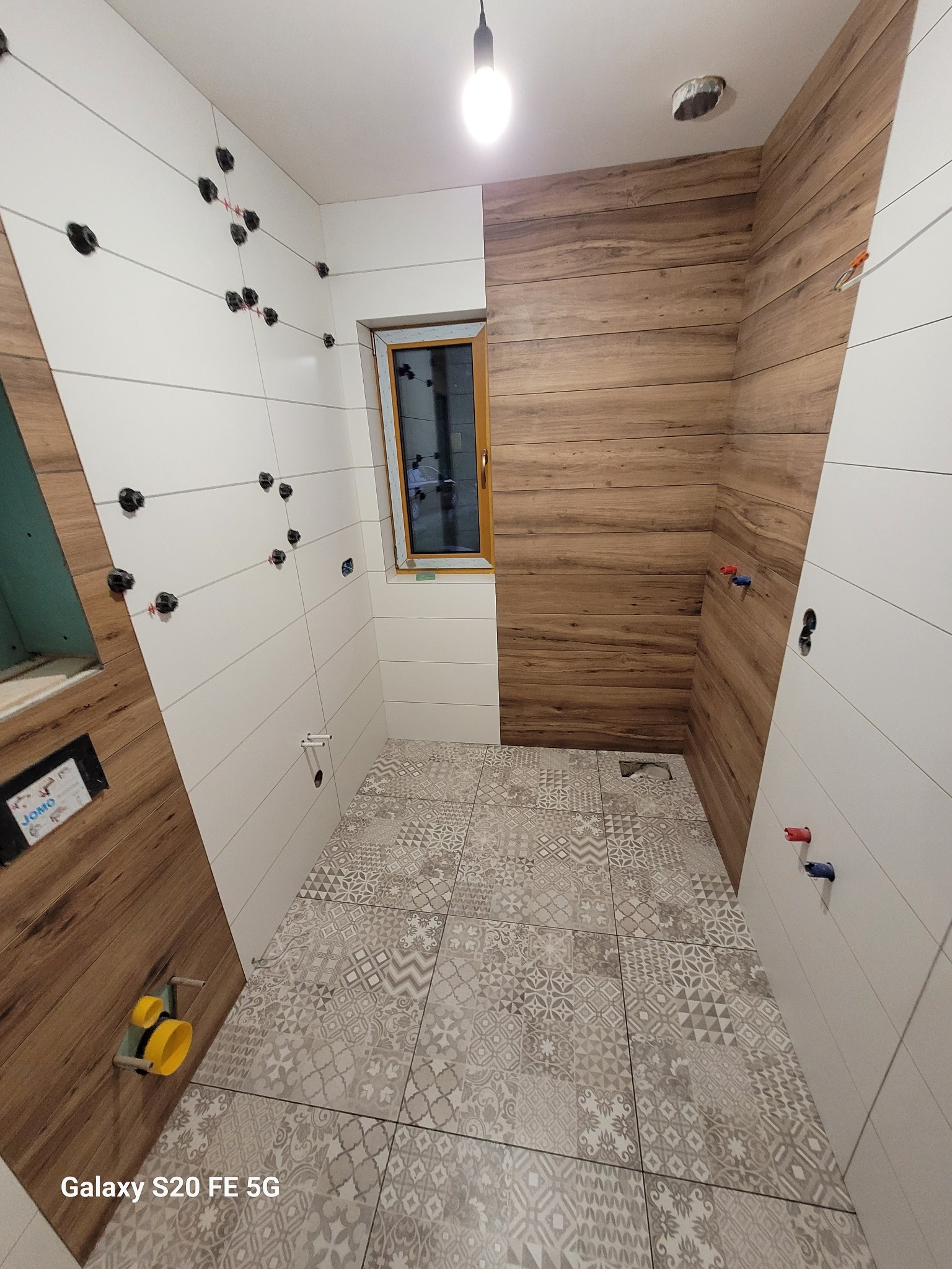 Łazienka w trakcie wykańczania: białe i drewnopodobne płytki ścienne ułożone naprzemiennie, wzorzysta szara podłoga, widoczne elementy instalacji wodnej i uchwyty do płytek na ścianach.