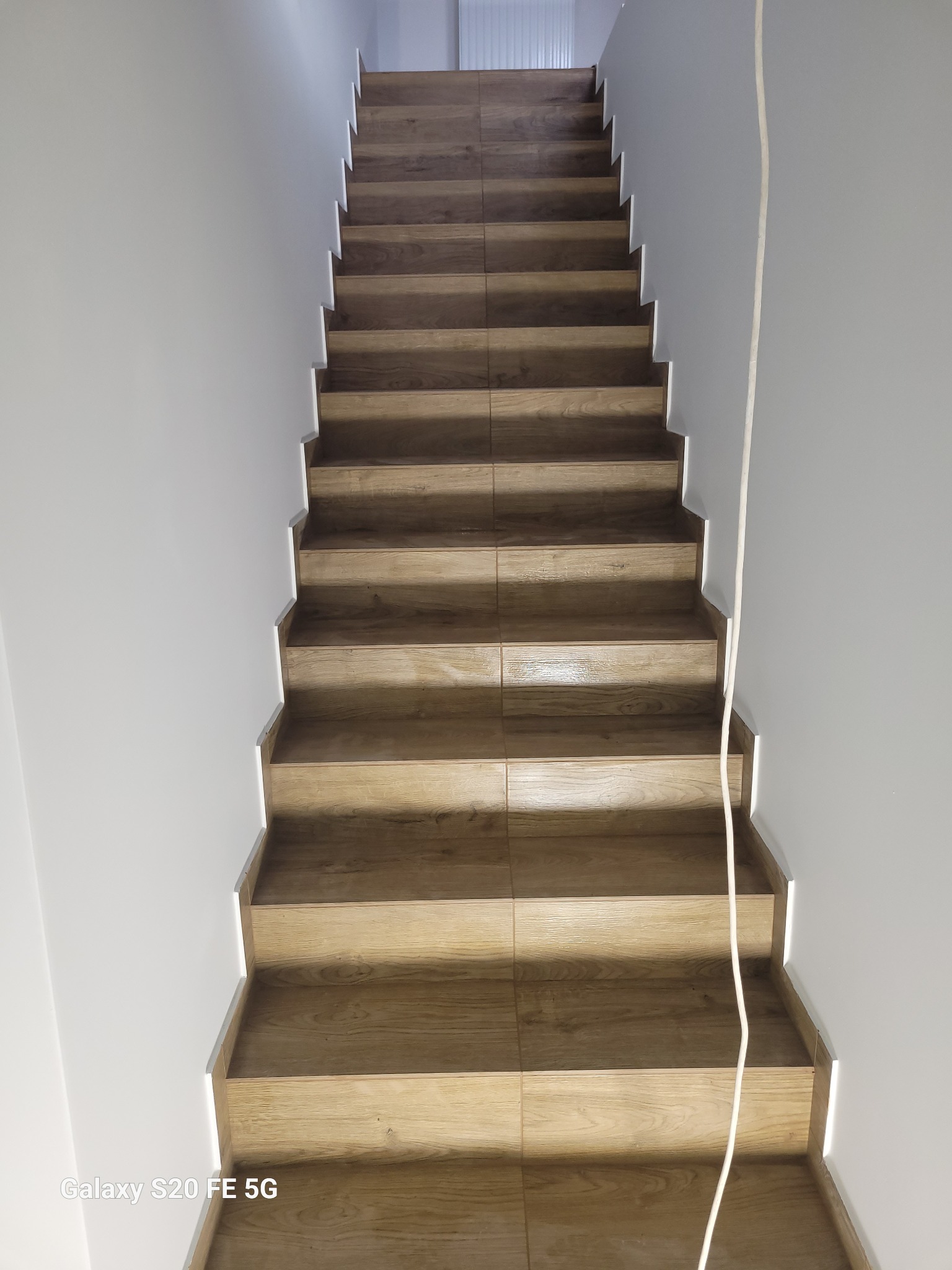 Schody wyłożone płytkami imitującymi drewno, prowadzące do góry, z białymi ścianami po bokach i widocznym kablem elektrycznym zwisającym z góry.