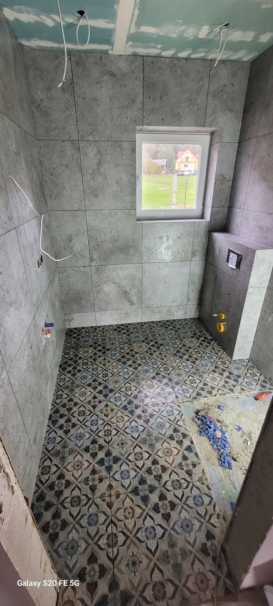 Wnętrze łazienki w trakcie prac: ułożona wzorzysta podłoga, szare płytki ścienne, widoczne instalacje, okno z widokiem na dom i zieleń.