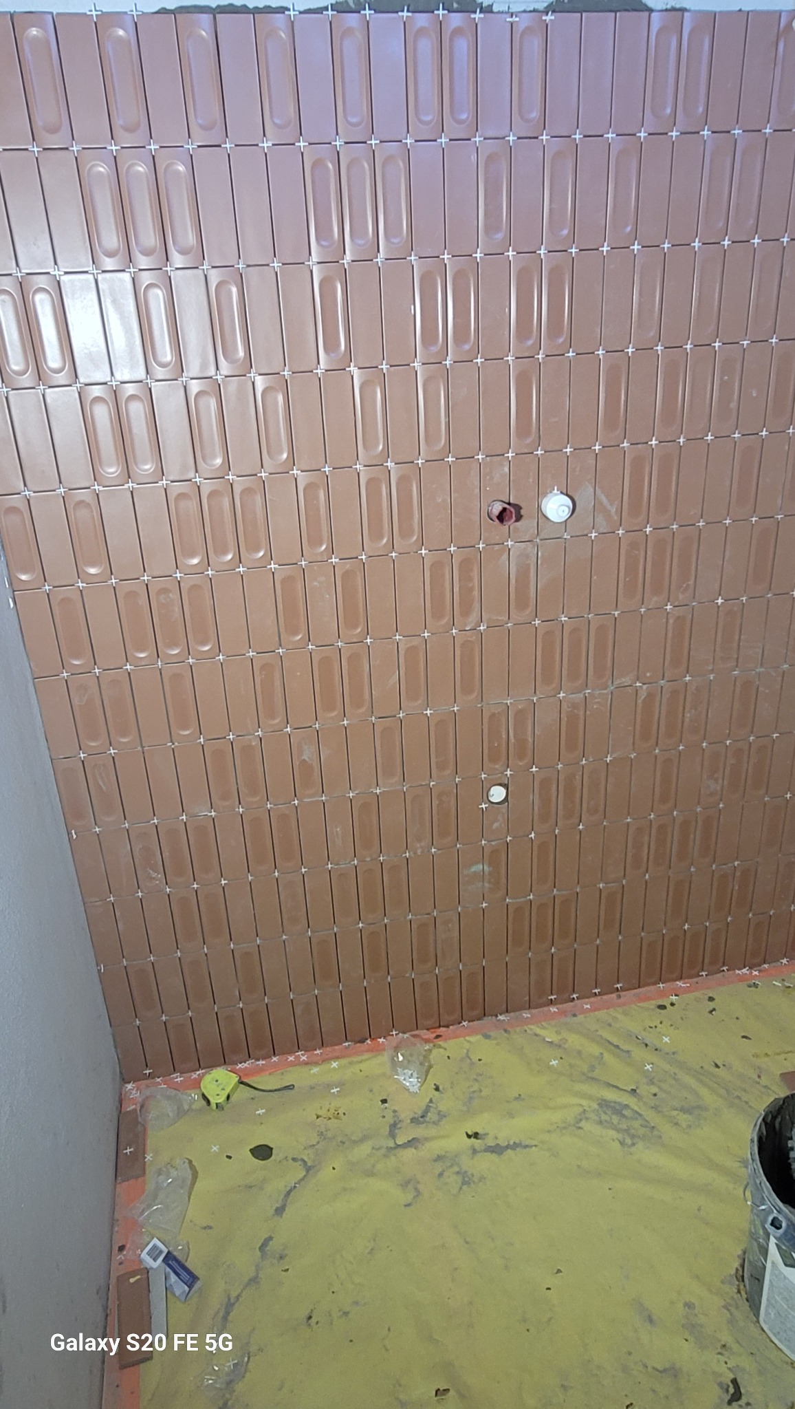 Układanie brązowych, podłużnych płytek na ścianie łazienki z użyciem krzyżyków dystansowych, widoczne przygotowane podłączenia hydrauliczne i zabezpieczona folią podłoga.