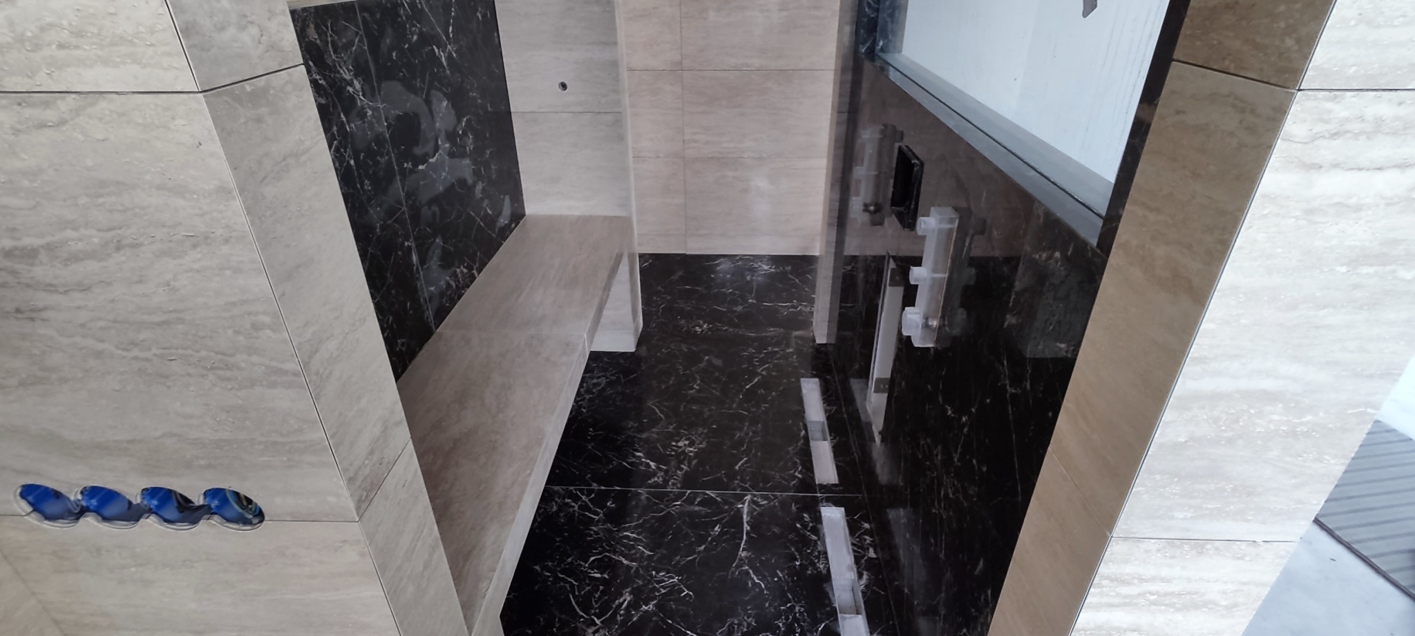 Nowoczesna łazienka z czarno-białymi marmurkowymi płytkami, beżowymi płytkami imitującymi trawertyn, wnęką z siedziskiem i odpływem liniowym pod kabiną prysznicową.