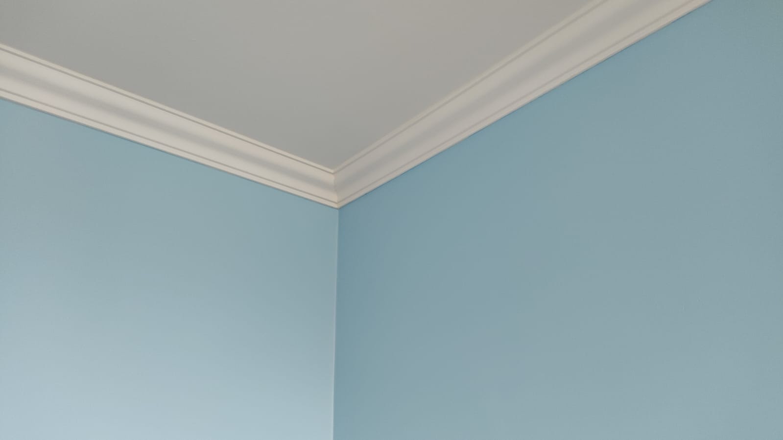 Ściana pomalowana na błękitno z białym, dekoracyjnym wykończeniem sufitu. Widoczny róg pokoju, gładka powierzchnia ściany i precyzyjne połączenie elementów.