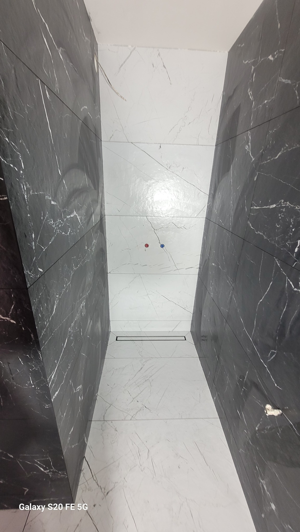 Wnętrze łazienki z czarno-białymi płytkami imitującymi marmur, ułożonymi wzdłuż ścian i na podłodze. Widoczny odpływ liniowy i wystające rurki.