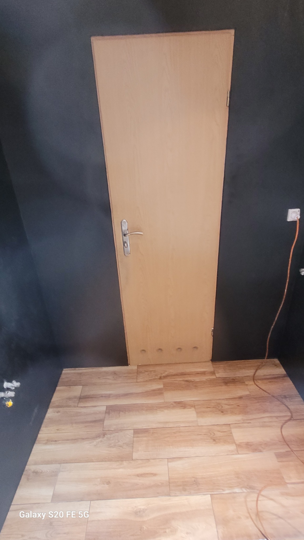 Jasne panele podłogowe z drewnianym wzorem, ściany pomalowane na ciemny kolor, widoczna drewniana futryna drzwiowa z metalową klamką i gniazdko elektryczne.