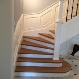 Klasyczne schody z drewnianymi stopniami i białymi podstopnicami, ozdobna biała balustrada, w tle marmurowa podłoga i koń na biegunach.