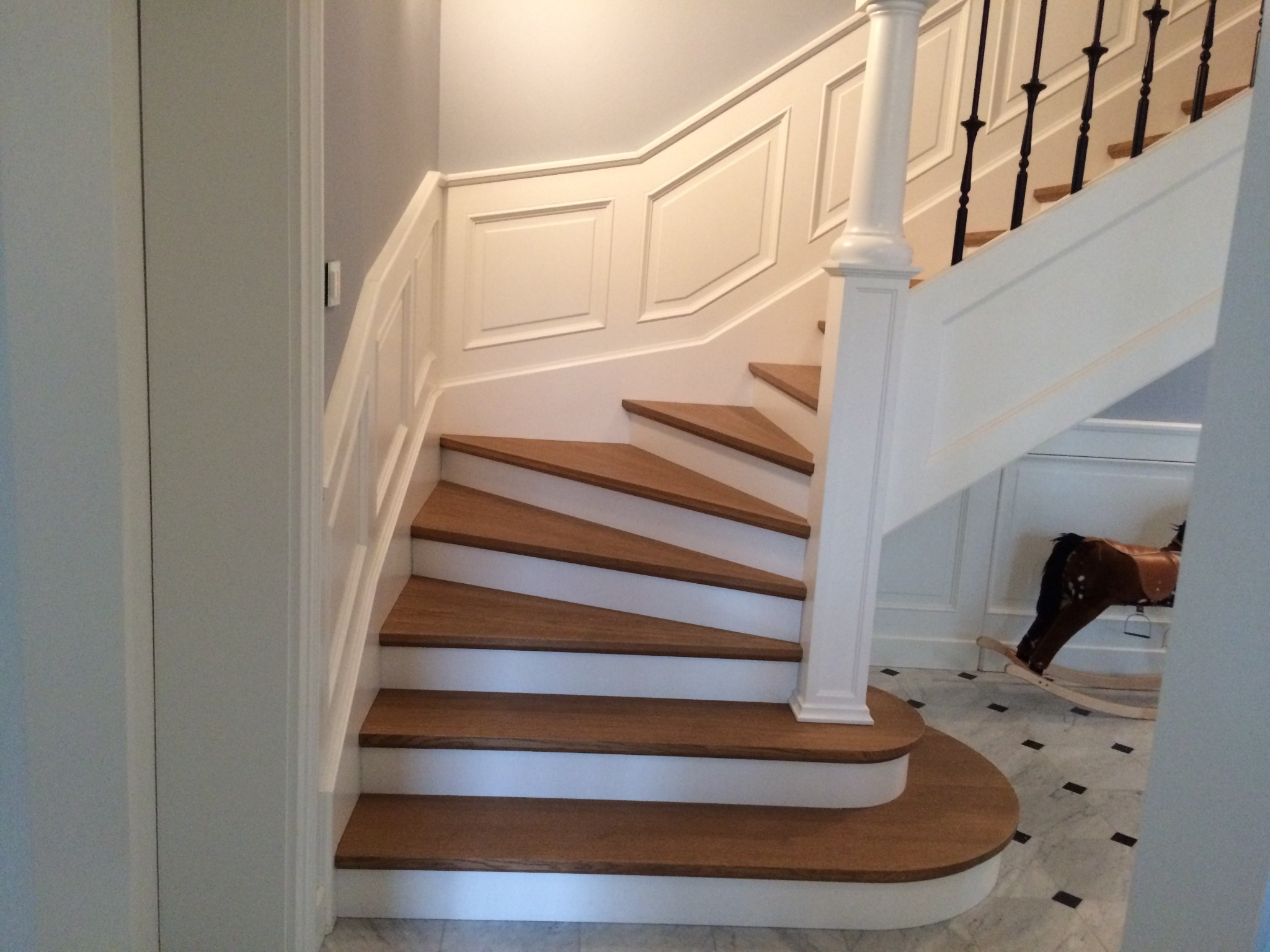 Klasyczne schody z drewnianymi stopniami i białymi podstopnicami, ozdobna biała balustrada, w tle marmurowa podłoga i koń na biegunach.