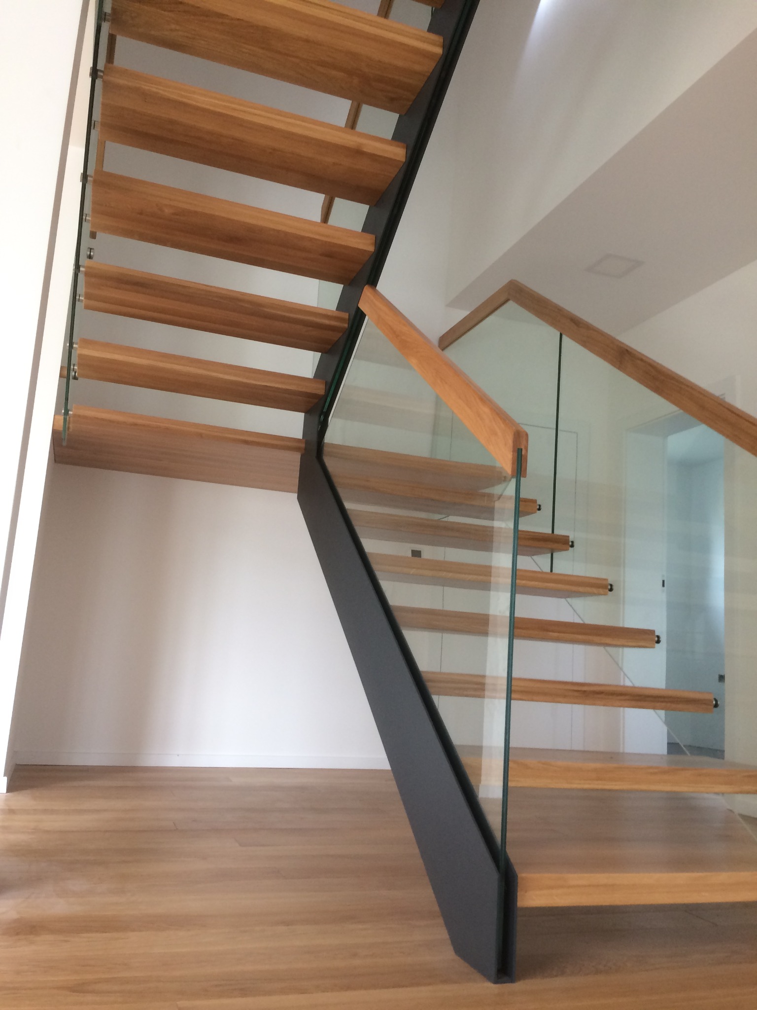 Nowoczesne schody z drewnianymi stopniami, szklanymi balustradami i czarną metalową konstrukcją, widok z dołu na jasnym parkiecie.