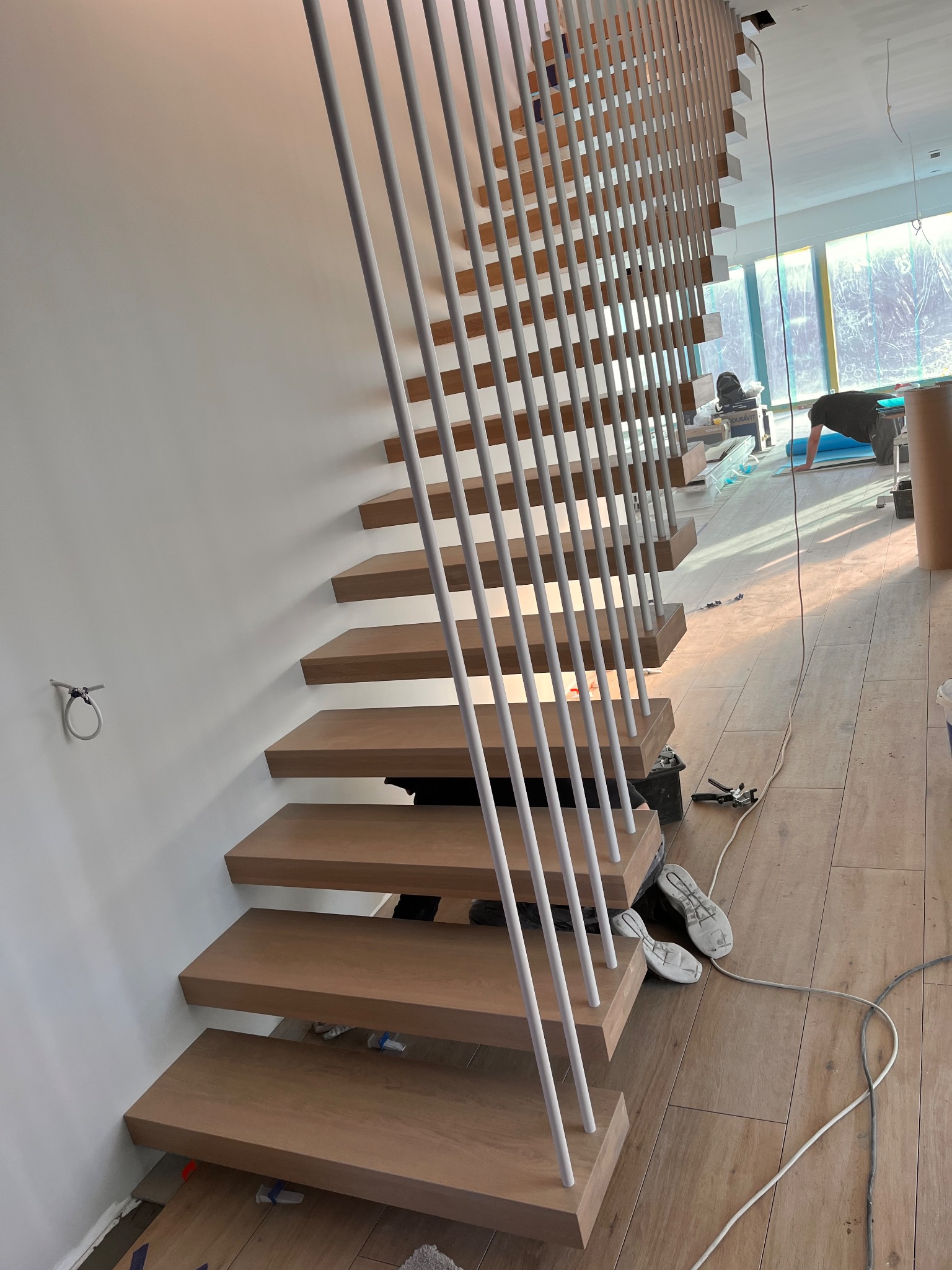 Nowoczesne, minimalistyczne schody z drewnianymi stopniami i białymi, pionowymi balustradami w trakcie montażu, widoczne narzędzia i osoba pracująca w tle.