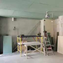 Wnętrze w trakcie montażu płyt gipsowo-kartonowych na suficie, widoczne rusztowanie, drabina, płyty oraz surowe ściany z instalacjami.