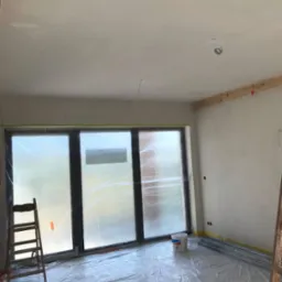 Przygotowanie do malowania: pomieszczenie zabezpieczone folią malarską, ściany częściowo wygładzone, drabina oparta o okno, widoczne puszki z farbą.