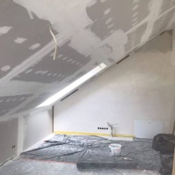 Denys Volos - Poddasze w trakcie prac wykończeniowych: widoczne płyty karton-gips na skosach sufitu, częściowo pokryte gładzią, zabezpieczona folią podłoga, puszka z zaprawą i narzędzia w pomieszczeniu z oknem...