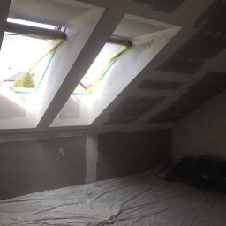 Denys Volos - Poddasze w trakcie prac wykończeniowych: ściany przygotowane pod malowanie, okna dachowe z zabezpieczeniem taśmą, podłoga przykryta folią ochronną, widoczne worki z materiałem.