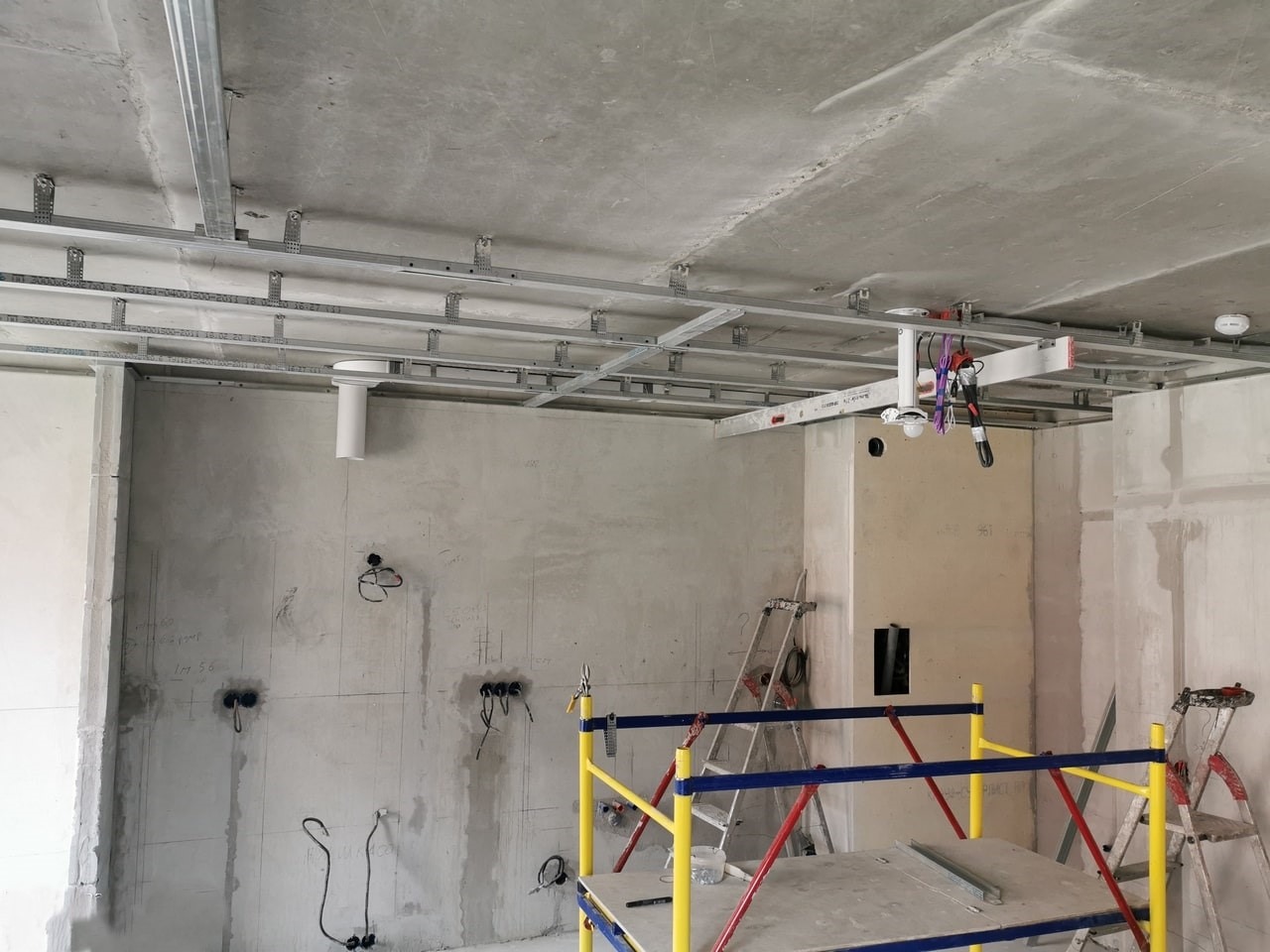 Montaż stelaża sufitu podwieszanego z płyt karton-gips, widoczne profile metalowe, przewody elektryczne i drabiny w surowym wnętrzu przed wykończeniem.