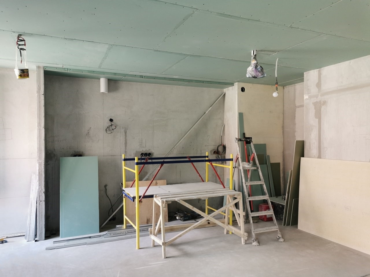 Wnętrze w trakcie montażu płyt gipsowo-kartonowych na suficie, widoczne rusztowanie, drabina, płyty oraz surowe ściany z instalacjami.