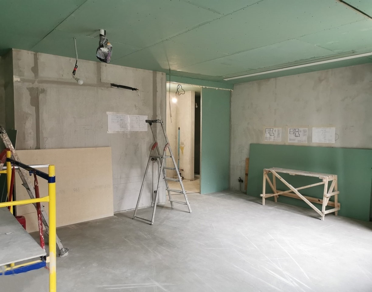 Wnętrze w trakcie prac wykończeniowych: surowy beton na ścianach, częściowo zamontowane płyty gipsowo-kartonowe na suficie i ścianach, drabiny, drewniany kozioł i plany architektoniczne na ścianie.