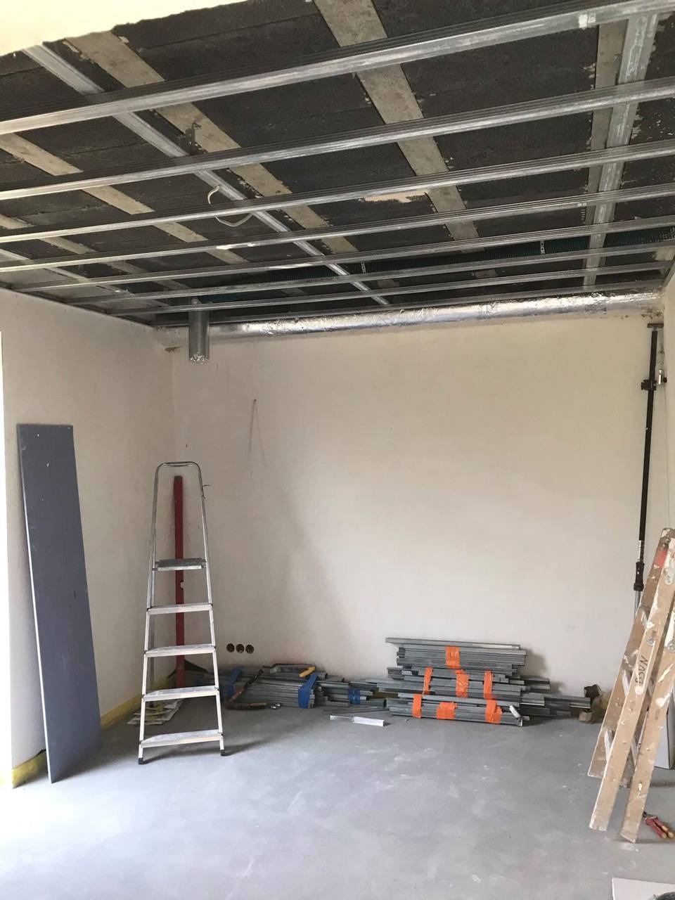 Konstrukcja metalowa sufitu podwieszanego z izolacją, widoczne profile, płyta karton-gips oparta o ścianę, drabiny i materiały budowlane w surowym wnętrzu.