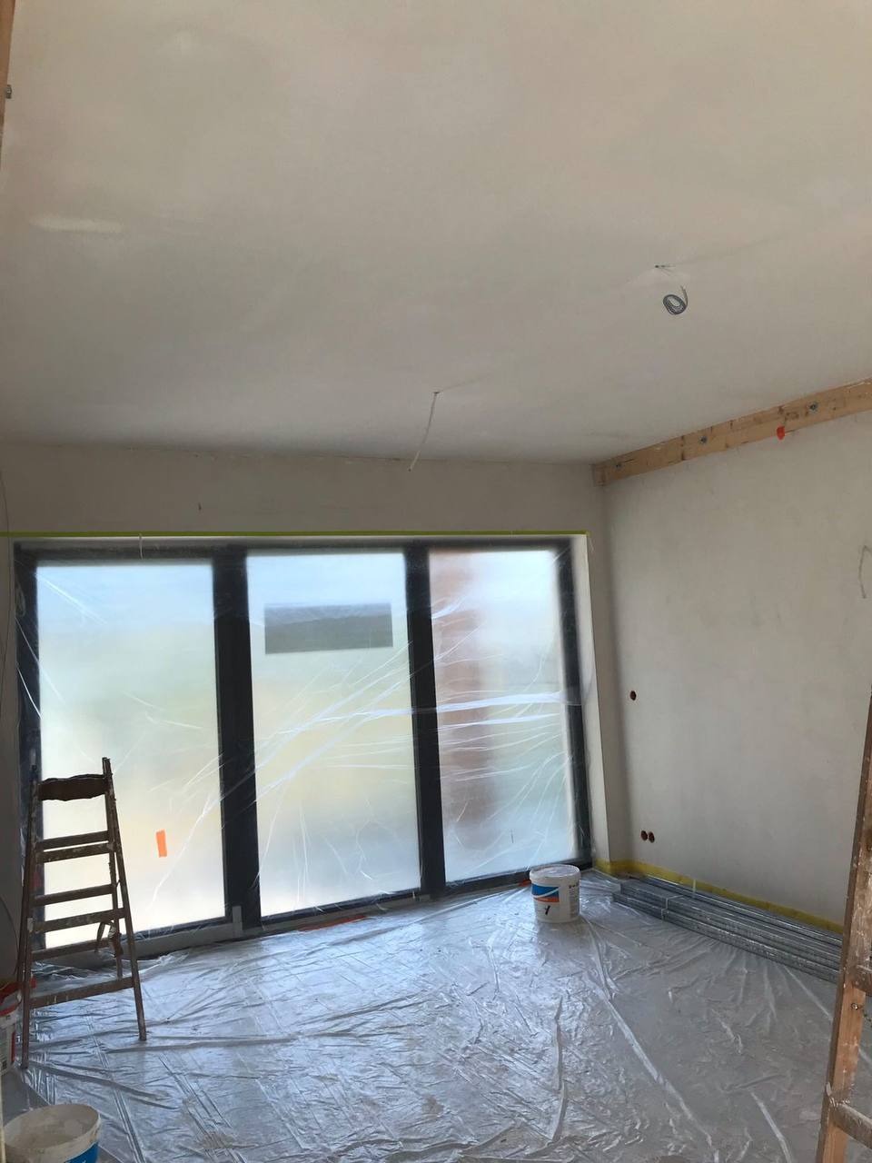 Przygotowanie do malowania: pomieszczenie zabezpieczone folią malarską, ściany częściowo wygładzone, drabina oparta o okno, widoczne puszki z farbą.