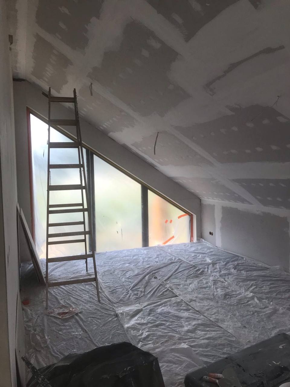 Poddasze w trakcie prac wykończeniowych: ściany i skośny sufit pokryte warstwą gipsu, okno zabezpieczone folią, podłoga przykryta plandeką ochronną, drewniana drabina oparta o ścianę.