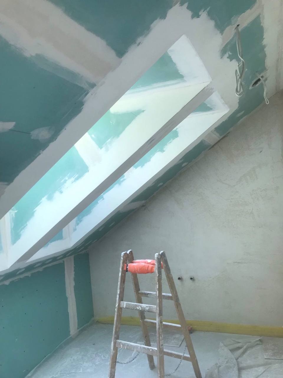 Poddasze w trakcie montażu płyt kartonowo-gipsowych, widoczna drewniana drabina, surowe ściany i fragment okna dachowego z obróbką.