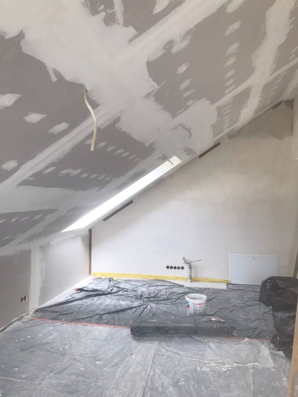 Poddasze w trakcie prac wykończeniowych: widoczne płyty karton-gips na skosach sufitu, częściowo pokryte gładzią, zabezpieczona folią podłoga, puszka z zaprawą i narzędzia w pomieszczeniu z oknem...