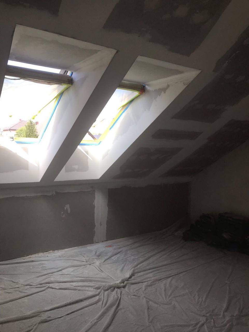 Poddasze w trakcie prac wykończeniowych: ściany przygotowane pod malowanie, okna dachowe z zabezpieczeniem taśmą, podłoga przykryta folią ochronną, widoczne worki z materiałem.