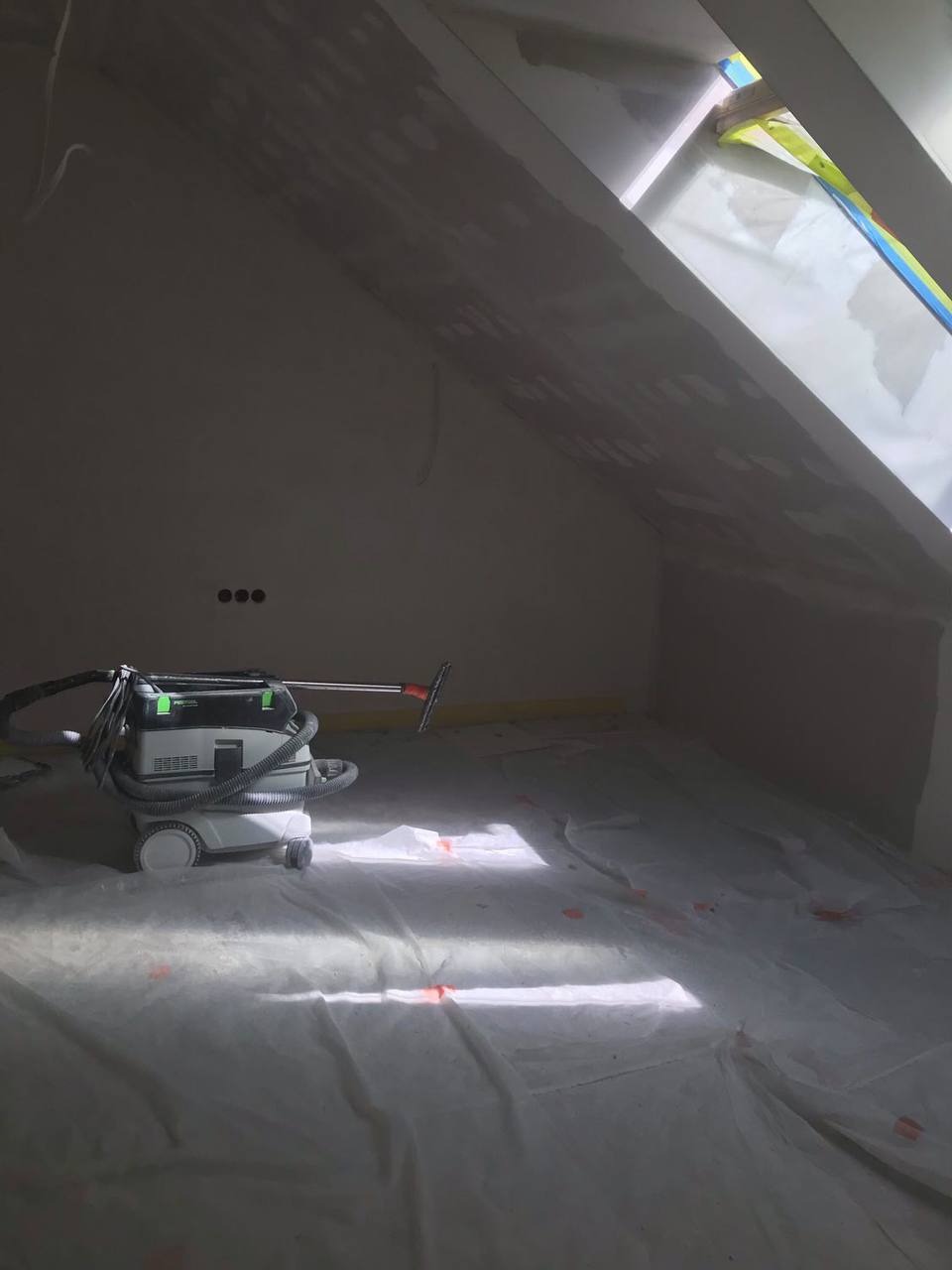 Poddasze w trakcie prac wykończeniowych, zabezpieczona folią podłoga, widoczny odkurzacz przemysłowy Festool, ściany przygotowane pod malowanie z widocznymi punktowymi nierównościami.
