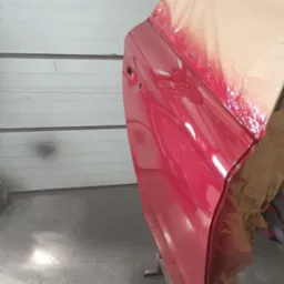 Czerwone drzwi samochodu częściowo oklejone papierem maskującym, przygotowane do lakierowania, widoczne na tle białej, garażowej bramy segmentowej.