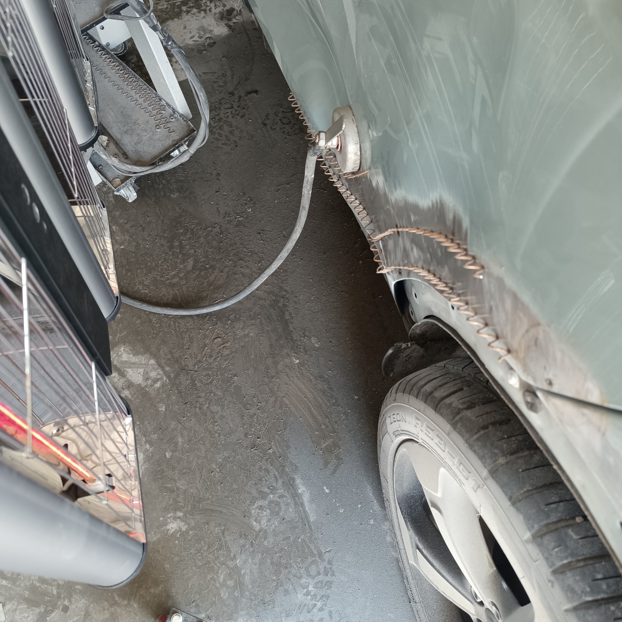 Przygotowanie nadkola samochodu do naprawy, z użyciem drutu do wyciągania wgnieceń, widoczna opona i fragment narzędzia blacharskiego.