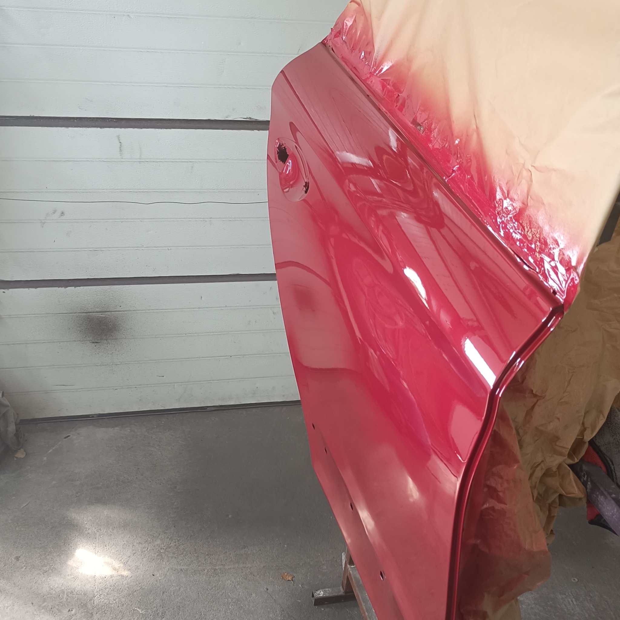 Czerwone drzwi samochodu częściowo oklejone papierem maskującym, przygotowane do lakierowania, widoczne na tle białej, garażowej bramy segmentowej.