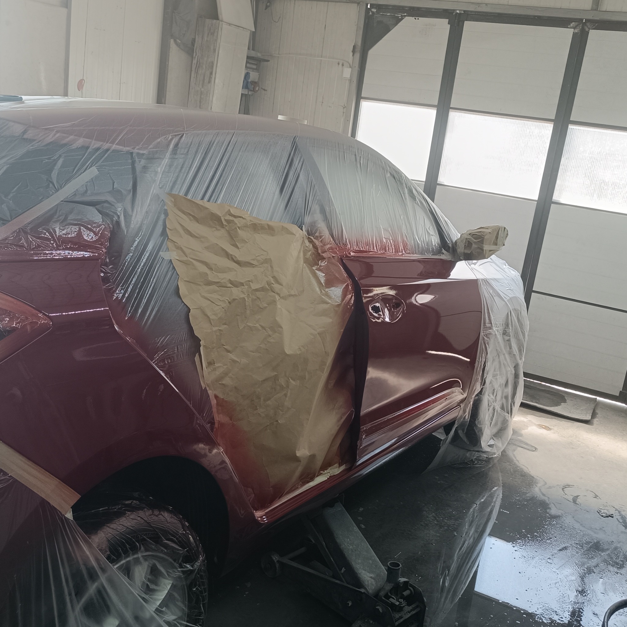Czerwony samochód w trakcie lakierowania, zabezpieczony folią i papierem maskującym, podniesiony na lewarku w warsztacie.