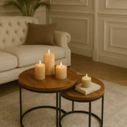 Zestaw dwóch okrągłych stolików kawowych z drewnianymi blatami i czarnymi metalowymi nogami, na których stoją zapalone świece w odcieniach beżu i zieleni, w tle kremowa sofa i dekoracyjna ściana.