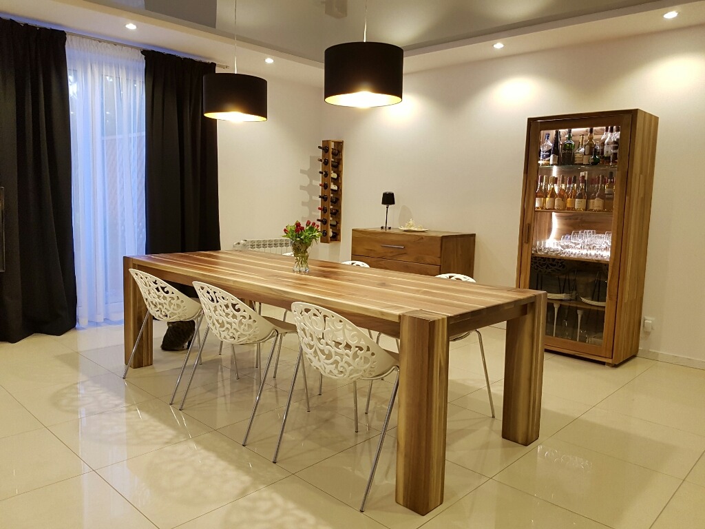 Jadalnia z dużym drewnianym stołem z litego drewna, białymi krzesłami o ażurowym wzorze, czarnymi lampami wiszącymi, komodą i witryną z oświetleniem LED, na podłodze z płytek w odcieniach beżu.