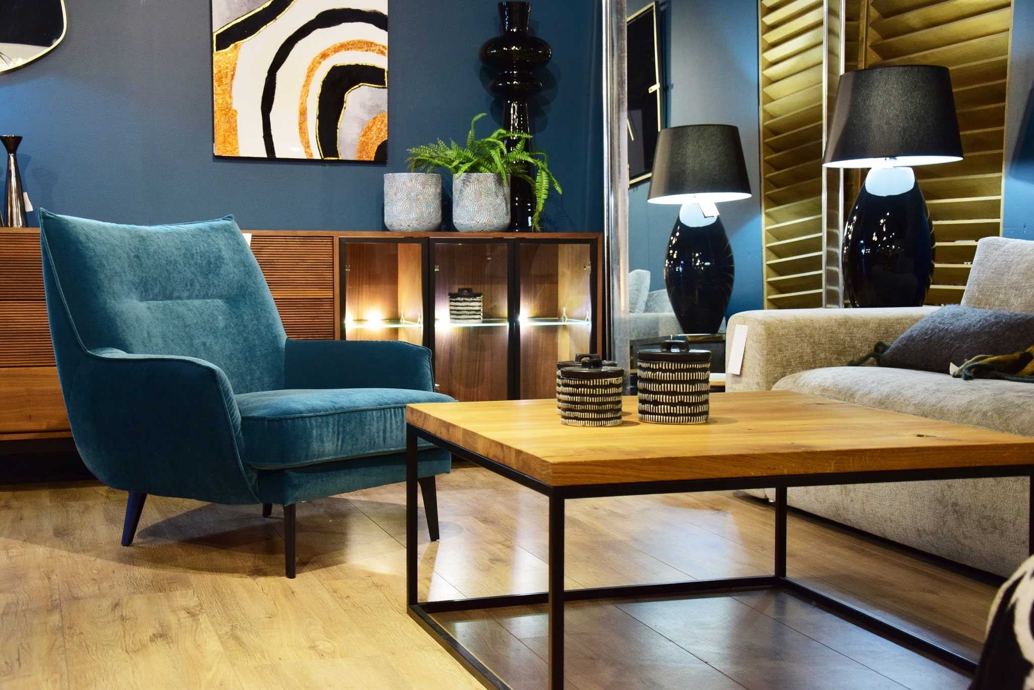 Salon z turkusowym fotelem klubowym, drewnianym blatem stolika kawowego na metalowej podstawie, drewnianą komodą z oświetleniem i dekoracjami.