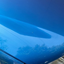 Scars Detailing - Błękitny lakierowany fragment karoserii samochodu z widocznymi smugami po polerowaniu, odbijający zieleń drzew. Widoczny fragment reflektora.