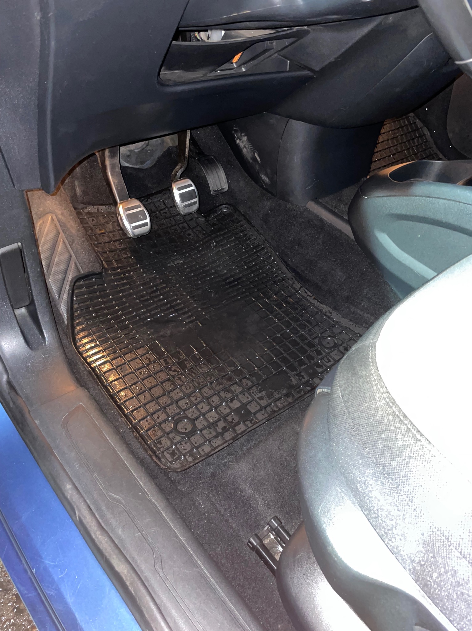 Wnętrze samochodu z gumową wycieraczką pod pedałami, na której widoczne są ślady wody lub wilgoci. Pedały z metalowymi nakładkami. Fragment fotela samochodowego po prawej stronie.