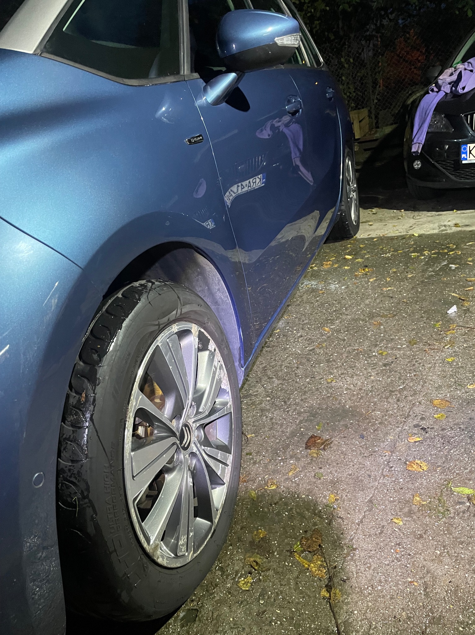 Błyszczący, błękitny samochód z widocznymi kroplami wody na oponie i karoserii, zaparkowany na mokrym, betonowym podłożu usianym liśćmi. Drugi, ciemny samochód widoczny w tle.