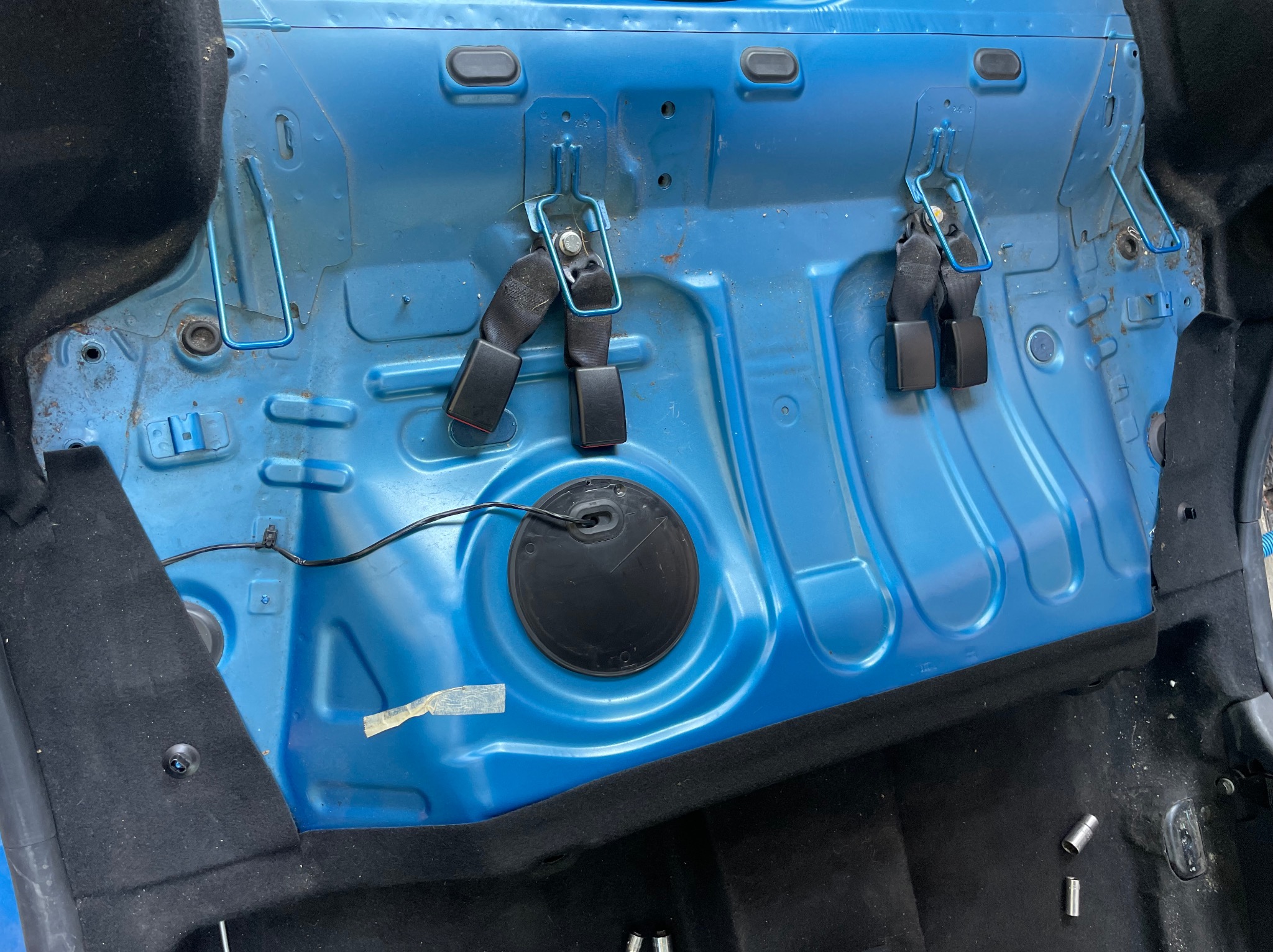 Wnętrze samochodu zdemontowane do czyszczenia, widoczne niebieskie elementy karoserii, czarne pasy bezpieczeństwa, przewody, oraz fragmenty czarnej tapicerki podłogi i tunelu środkowego.