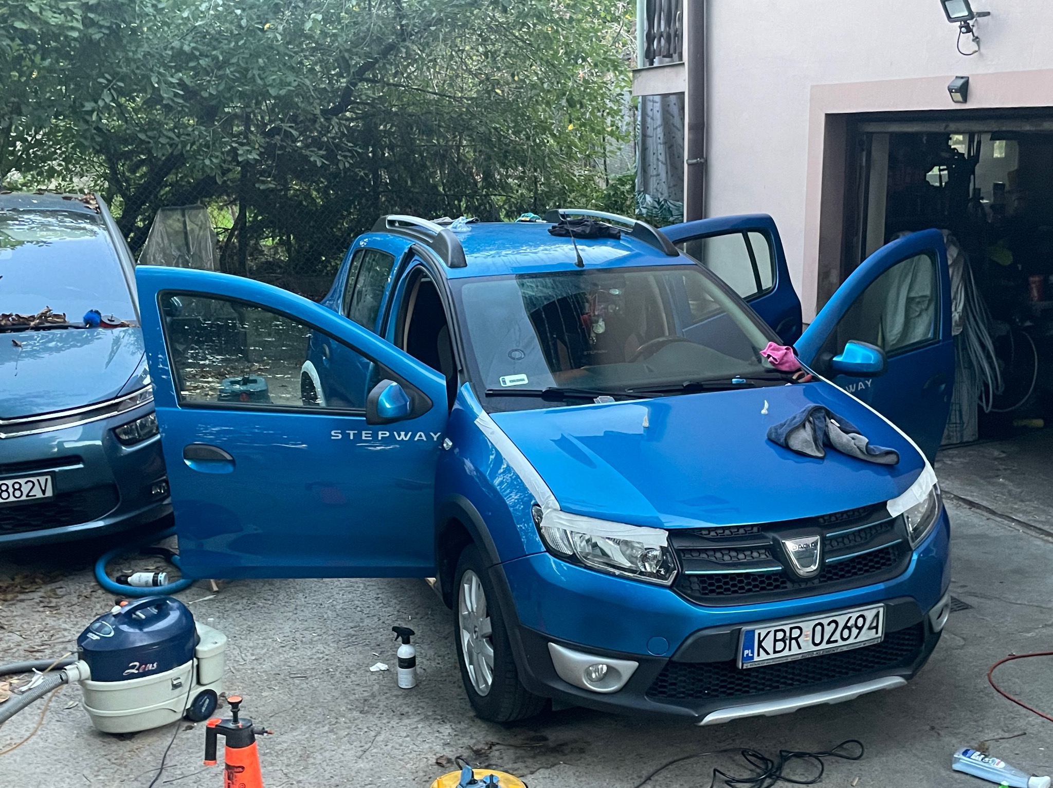 Niebieska Dacia Stepway z otwartymi drzwiami podczas czyszczenia; widoczny odkurzacz piorący i środki czyszczące, tablica rejestracyjna KBR 02694, częściowo widoczny drugi samochód i garaż w tle.