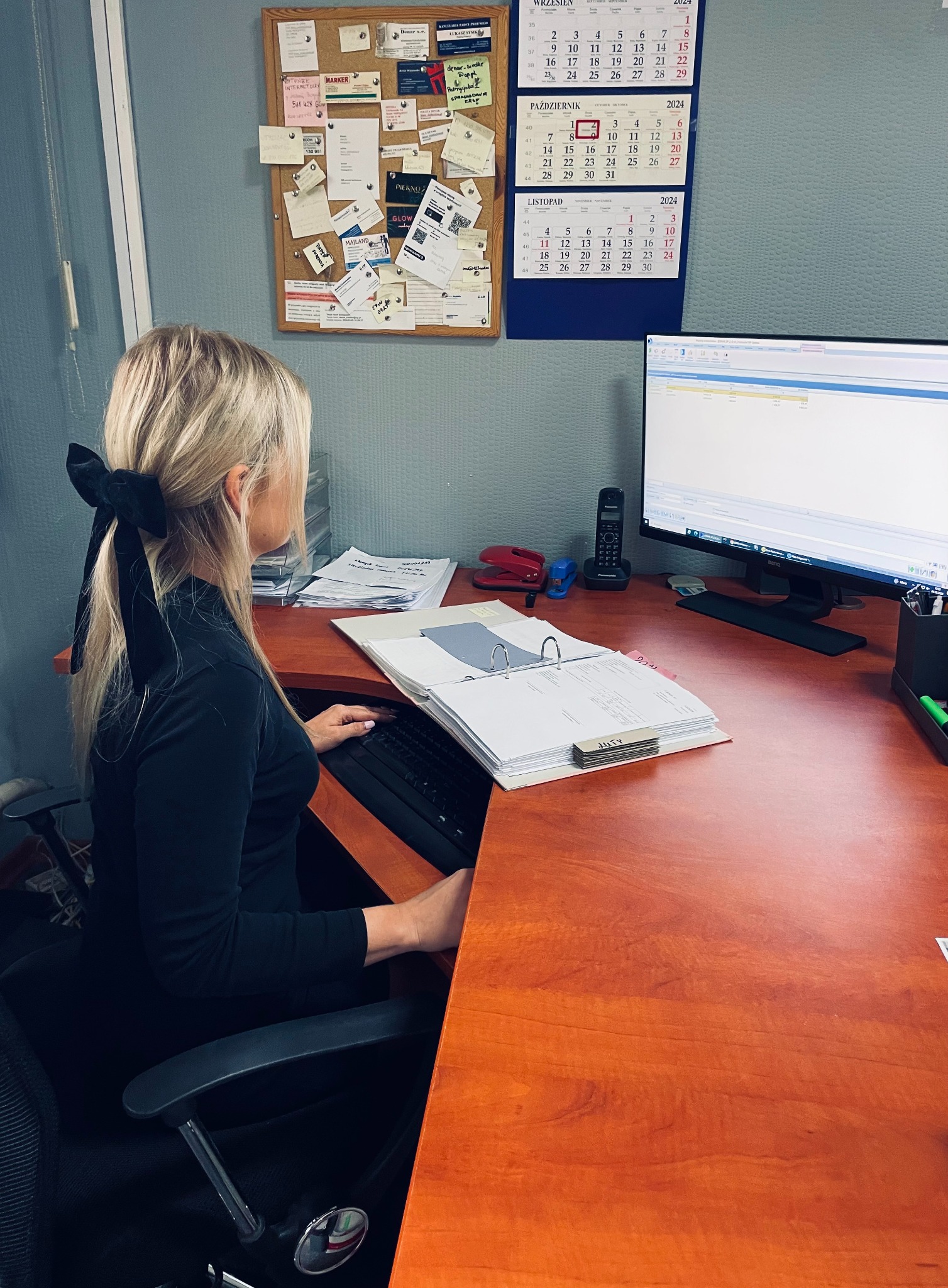 Kobieta z blond włosami spiętymi czarną kokardą, siedzi przy biurku z komputerem i segregatorami z dokumentami, w tle tablica korkowa z notatkami i kalendarze.