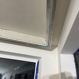 Kacper Klimek - Montaż płyty KG na suficie z widocznymi profilami, narożnikami i oknem w tle. Etap wykończenia wnętrza, przygotowanie do dalszych prac.