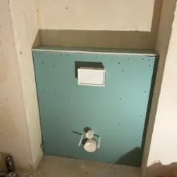 Zielona płyta GK z wyciętymi otworami na instalacje, zamontowana w narożniku pomieszczenia w trakcie prac wykończeniowych. Widoczne narzędzia.