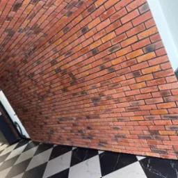 Ściana z cegieł dekoracyjnych w odcieniach czerwieni i brązu, ułożona pod kątem, z czarno-białą podłogą w szachownicę i widocznym oświetleniem sufitowym.