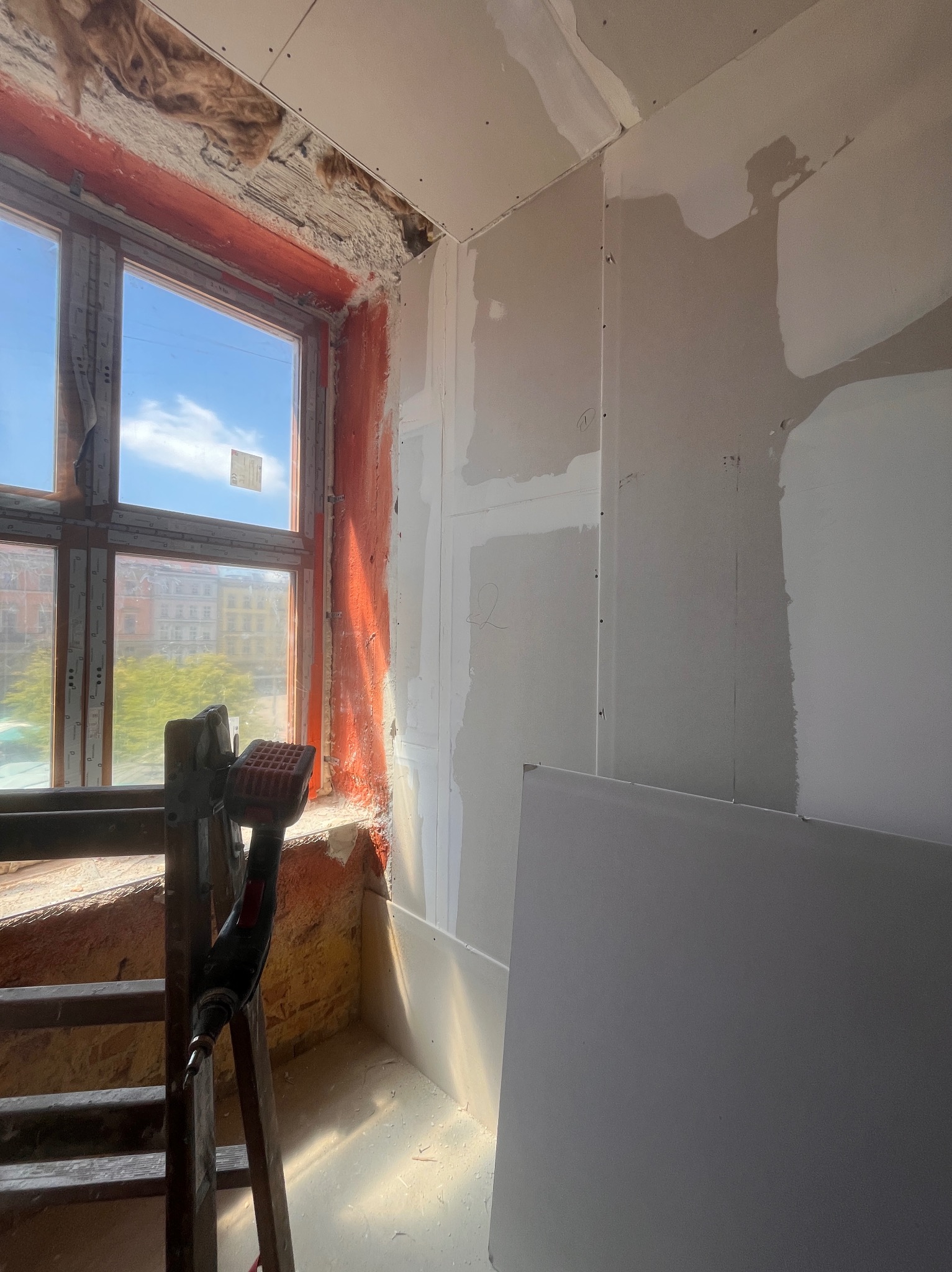Wnętrze w trakcie prac: zamontowane płyty gipsowe na ścianach, widoczne ocieplenie okna, drabina i wkrętarka. Dziennie światło wpada przez okno.